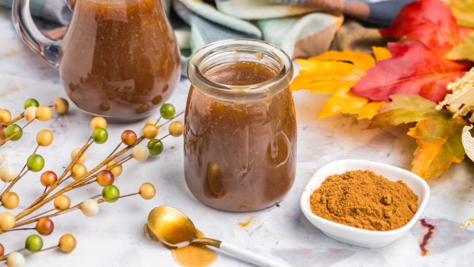 Jars of syrup beside a bowl of spices are surrounded by fall decorations.