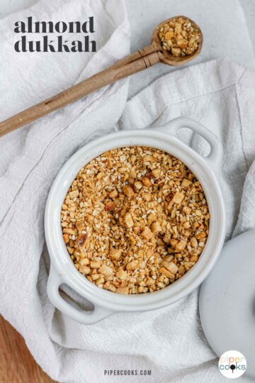 Close-up of homemade almond dukkah in a small bowl, showing toasted almonds, sesame seeds, and spices with the text almond dukkah.