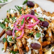 Close-up of golden Dukkah-coated fries with tahini yogurt sauce, feta, olives, and pickled red onions.