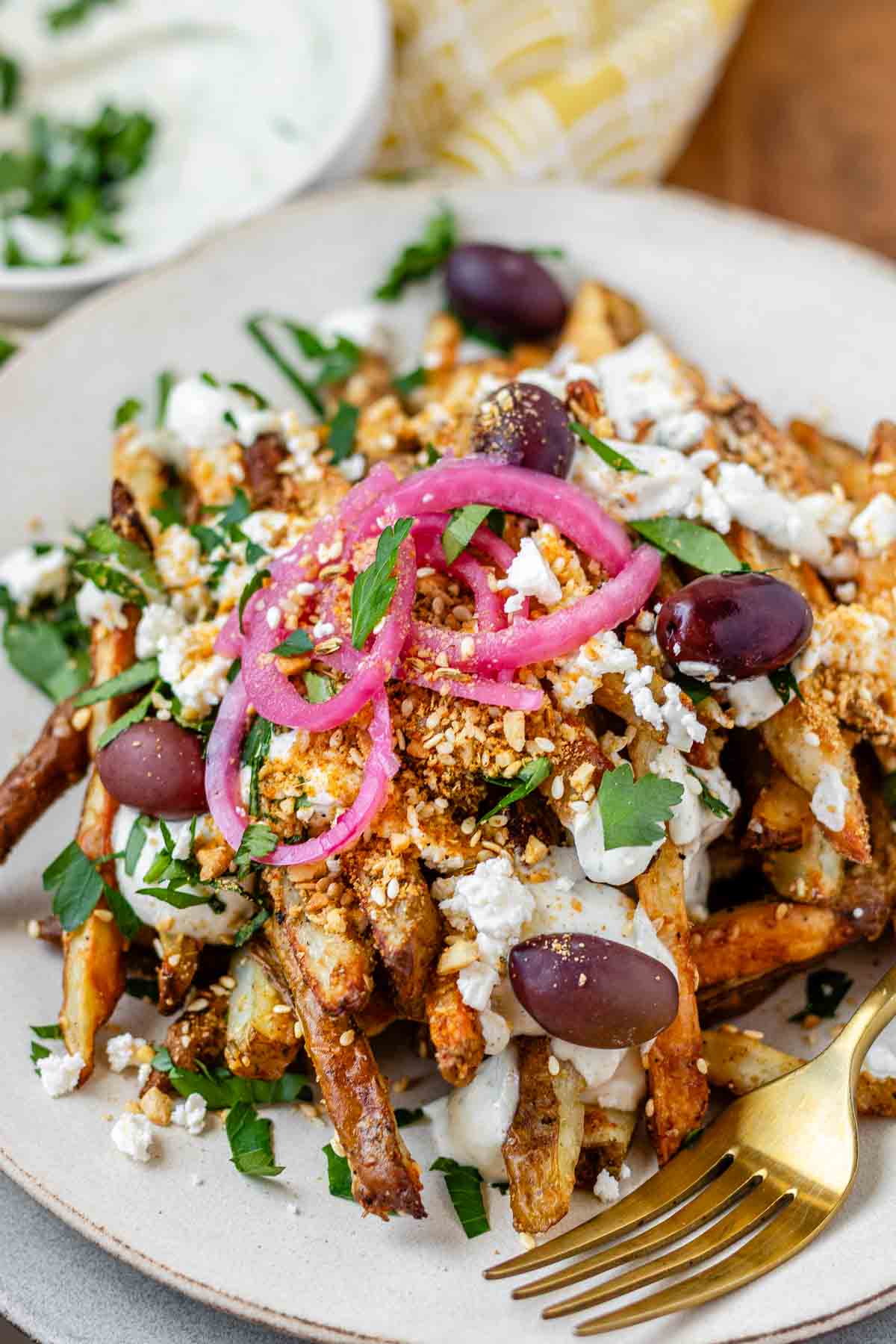 Close-up of golden Dukkah-coated fries with tahini yogurt sauce, feta, olives, and pickled red onions.