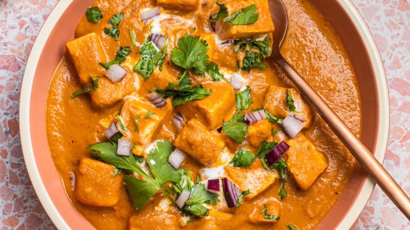 A bowl of paneer butter masala with red onion and herbs.