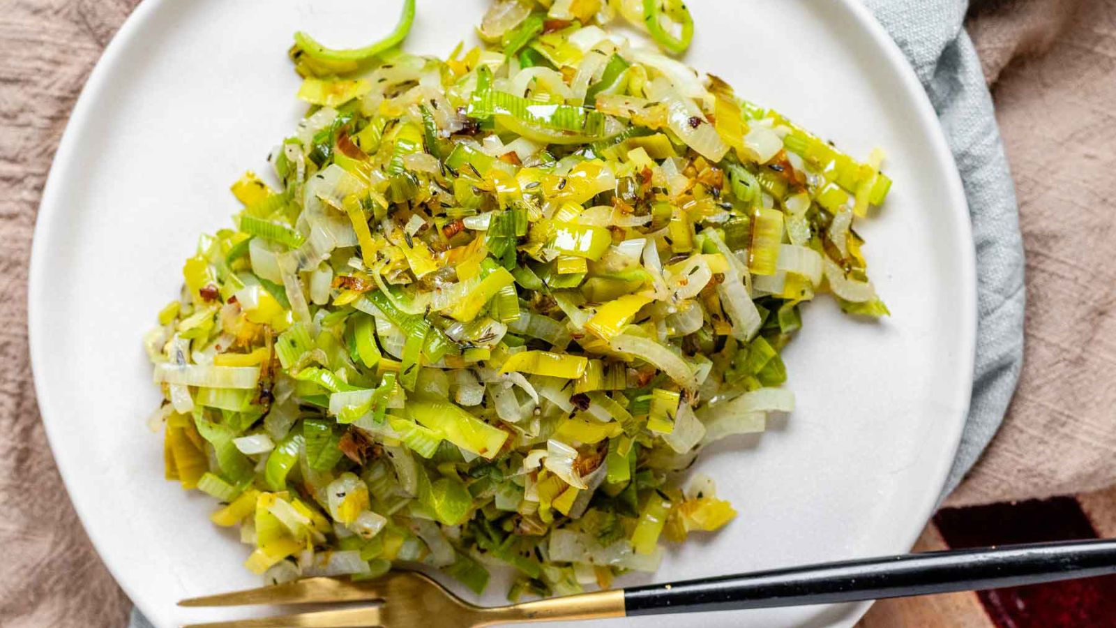 Chopped sauteed leeks on a white plate with a black and gold fork.