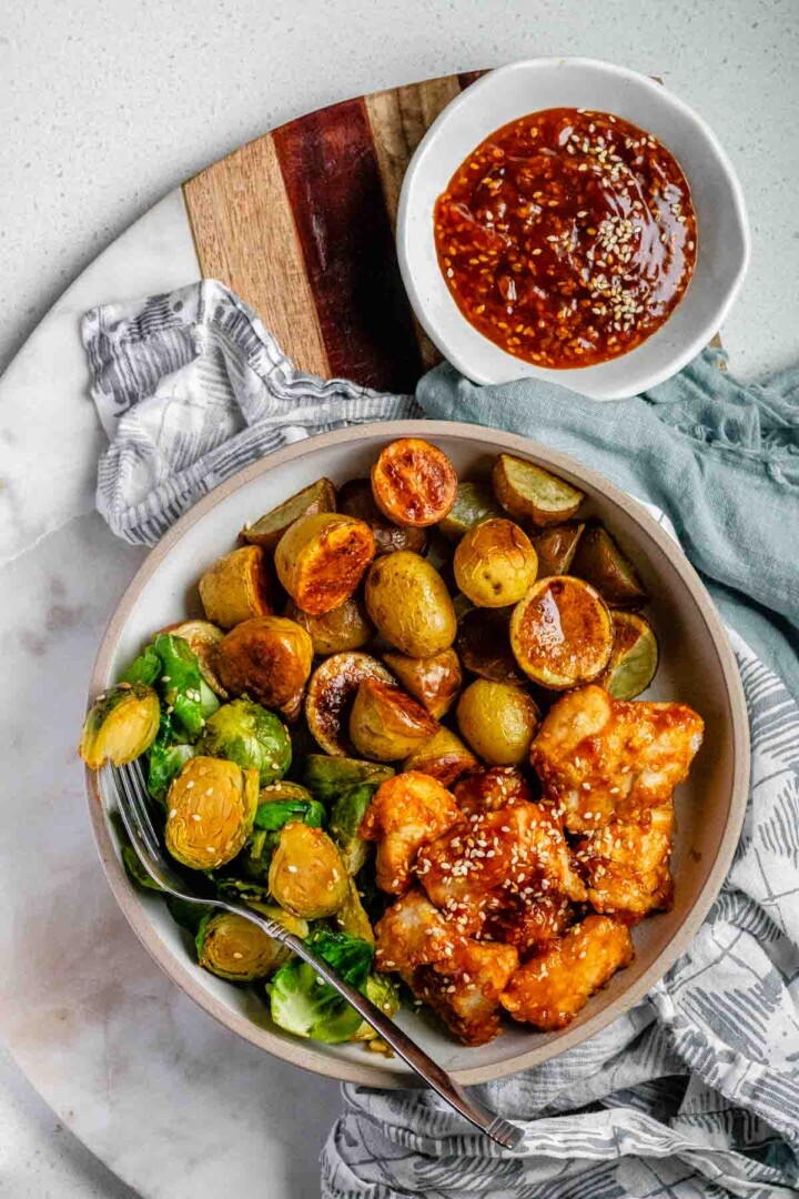 Plated serving of spicy sesame chicken, baby potatoes, and Brussels sprouts with extra sesame sauce on the side.