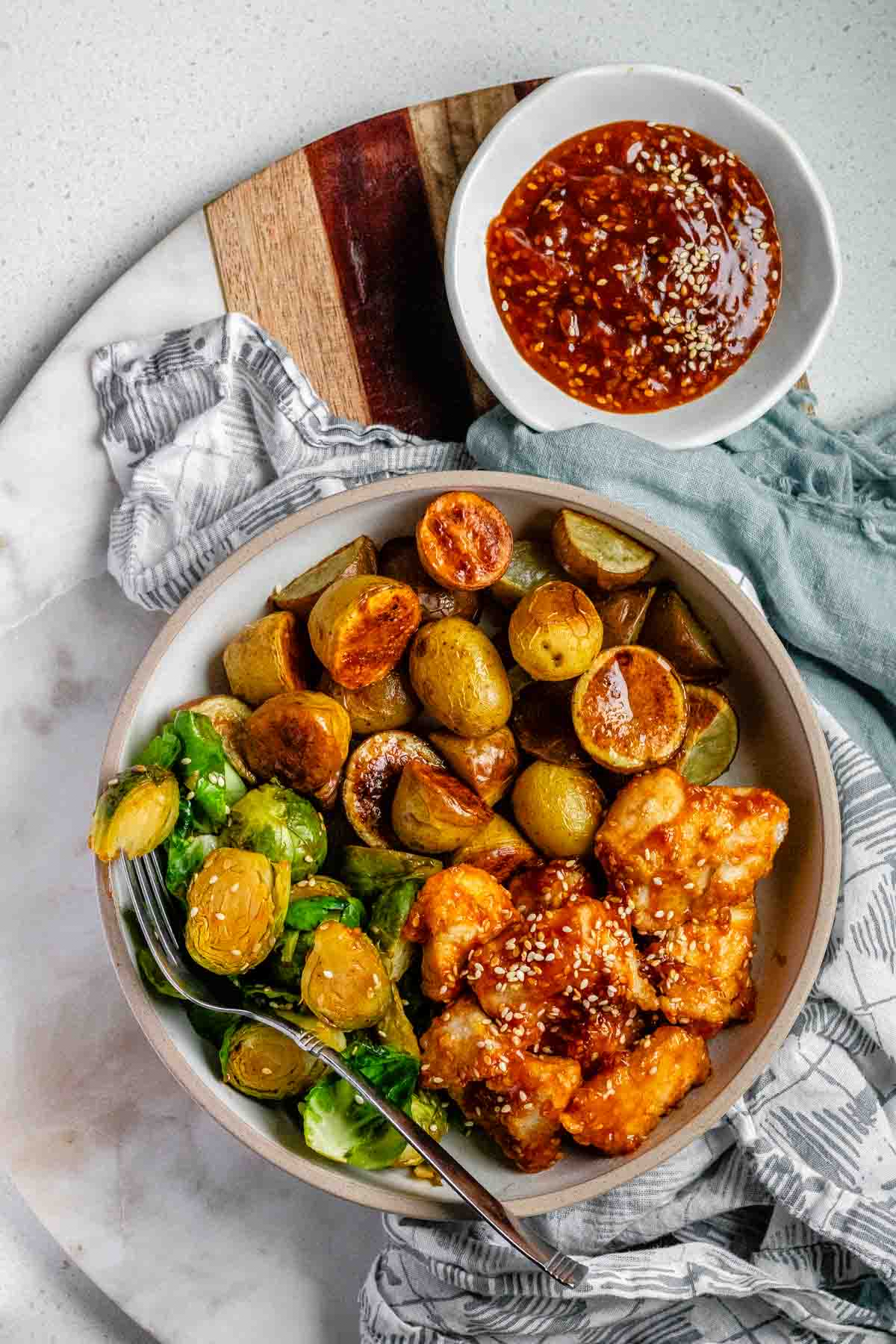 Plated serving of spicy sesame chicken, baby potatoes, and Brussels sprouts with extra sesame sauce on the side.