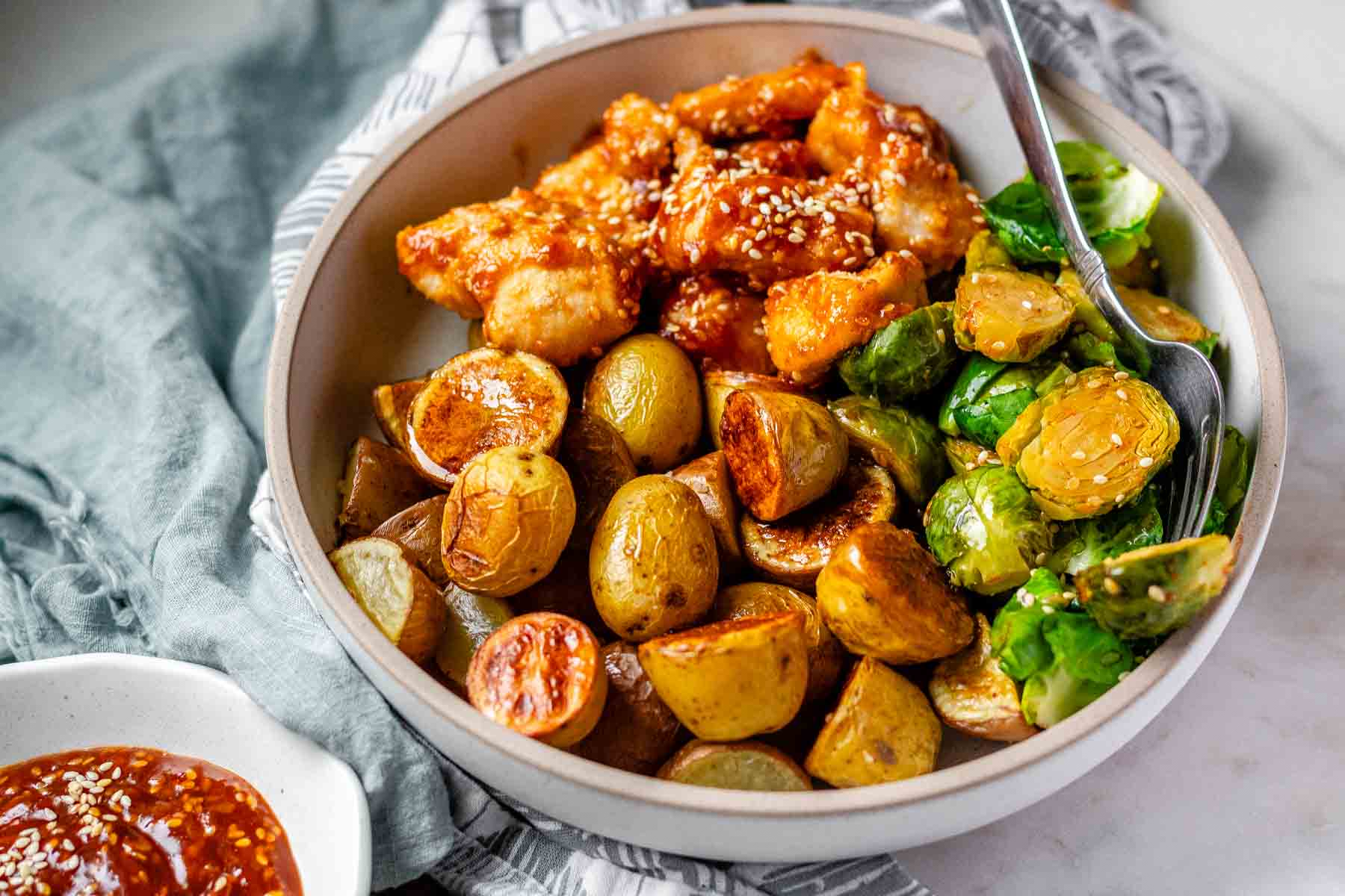 Plated serving of spicy sesame chicken, baby potatoes, and Brussels sprouts with extra sesame sauce on the side.