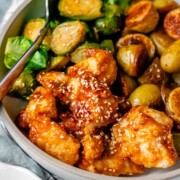 Plated serving of spicy sesame chicken, baby potatoes, and Brussels sprouts with extra sesame sauce on the side.