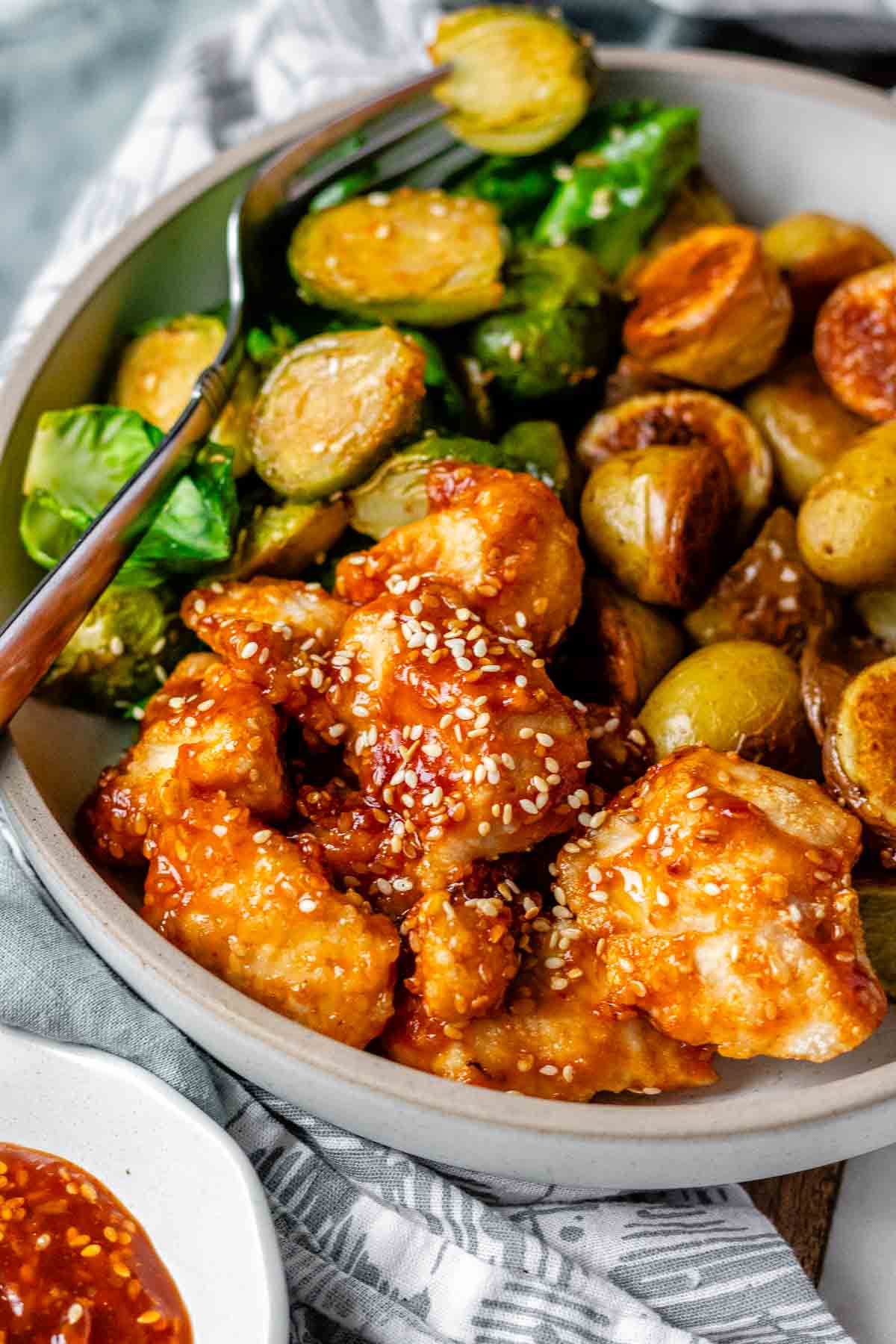 Plated serving of spicy sesame chicken, baby potatoes, and Brussels sprouts with extra sesame sauce on the side.