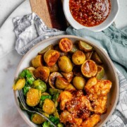 Plated serving of spicy sesame chicken, baby potatoes, and Brussels sprouts with extra sesame sauce on the side with text sheet pan chicken & veg.