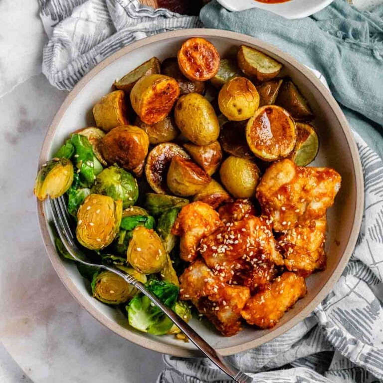 Plated serving of spicy sesame chicken, baby potatoes, and Brussels sprouts with extra sesame sauce on the side.