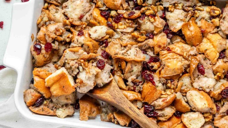 A close up of walnut cranberry stuffing in a white casserole dish with a wooden spoon.