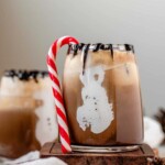 Mint chocolate iced coffee in a clear glass with marshmallow fluff snowmen visible on the inside of the glass, chocolate drizzle, and a candy cane.