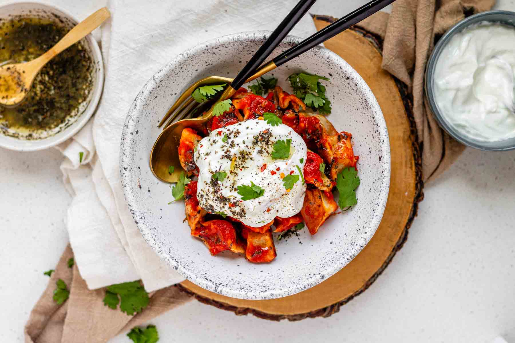 A bowl of dumplings in red sauce topped with yogurt and herbs.