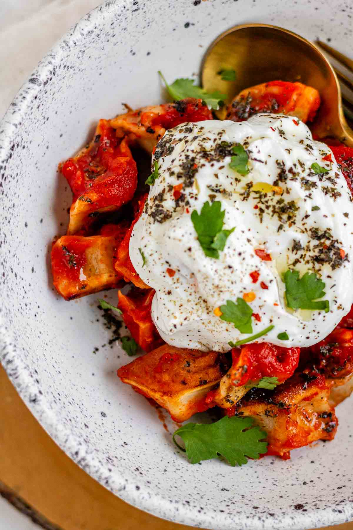 A bowl of dumplings in red sauce topped with yogurt and herbs.