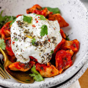 A bowl of dumplings in red sauce topped with yogurt and herbs with the text manti with beyond beef.