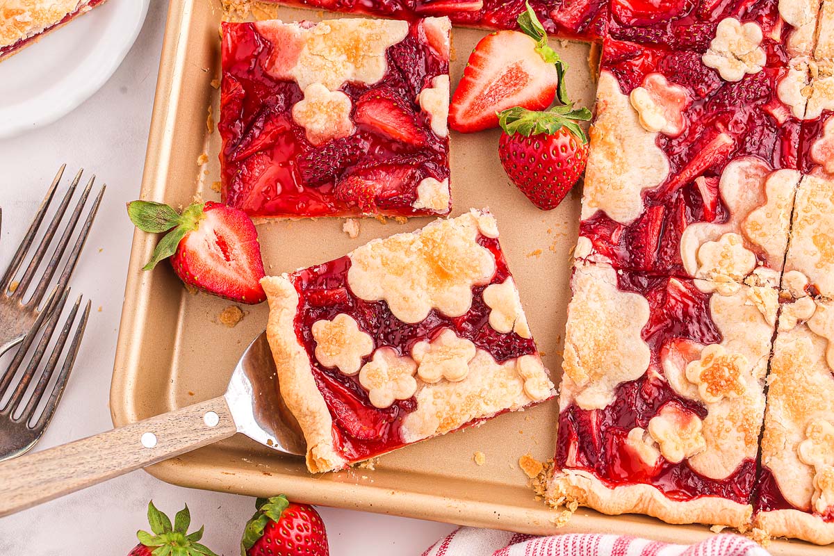 A baking tray with a strawberry slab pie, its square servings topped with flower-shaped crusts and surrounded by fresh strawberries.