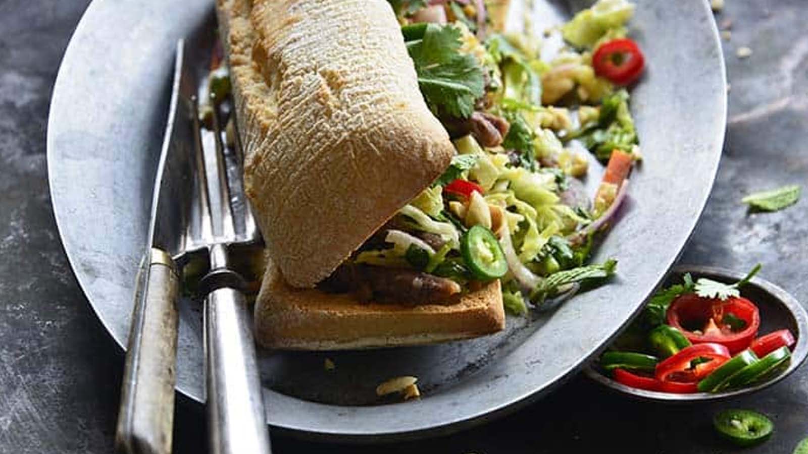 A sandwich loaded with meat and vegetables on a plate with a knife and fork.