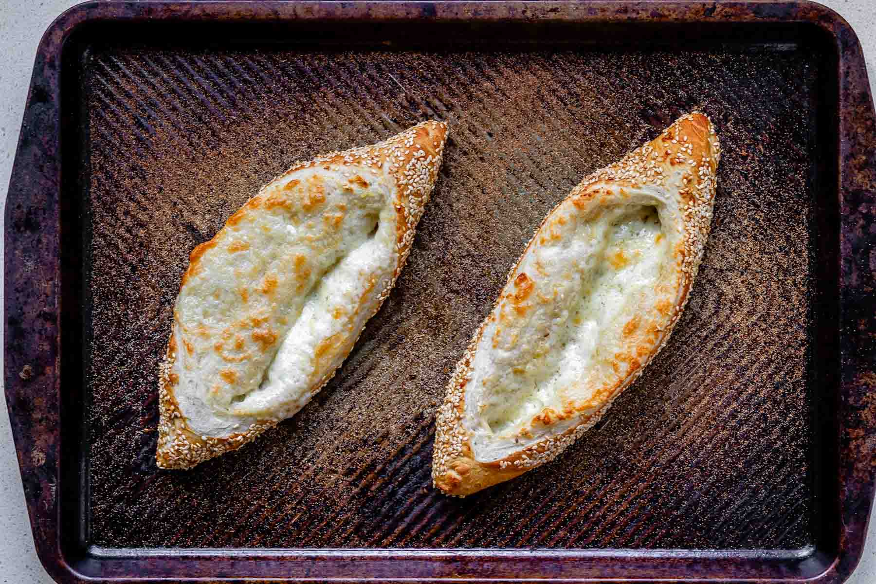 Boat-shaped baked sesame coated dough filled with melted cheese on a baking sheet.