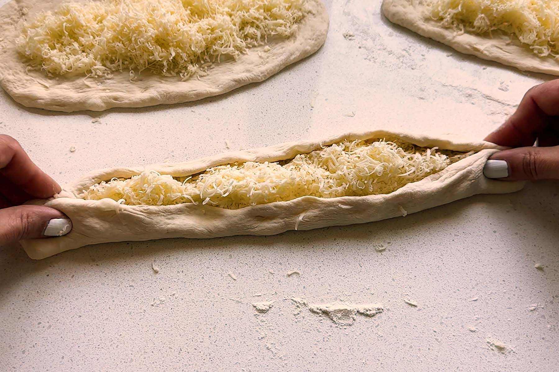 A long oval of cheese topped dough is pinched into a boat shape.