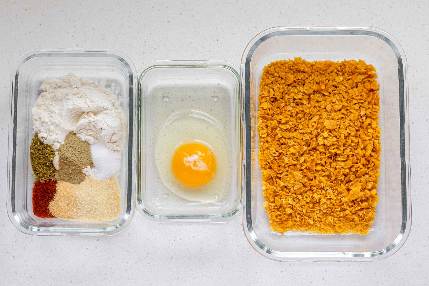 A breading station for chicken strips with a container of spices and flour, one with an egg, and one with crushed cornflakes.