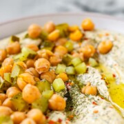 Hummus in a bowl topped with olive oil, chickpeas, chopped pickles, and spices.