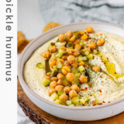 Hummus in a bowl topped with olive oil, chickpeas, chopped pickles, and spices with the text dill pickle hummus.