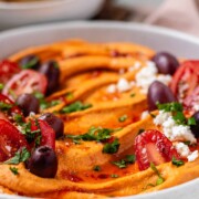 Orange hummus in a bowl topped with olive oil, crumbly white cheese, kalamata olives, chopped herbs, and halved tomatoes beside a bowl of crackers.