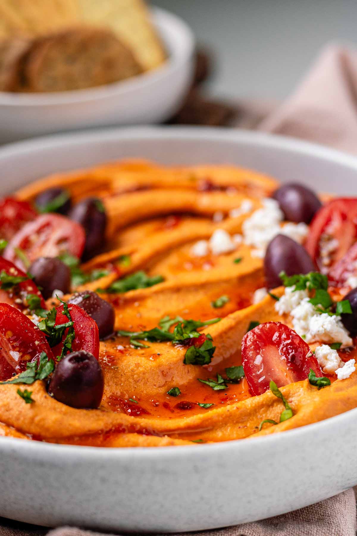Orange hummus in a bowl topped with olive oil, crumbly white cheese, kalamata olives, chopped herbs, and halved tomatoes beside a bowl of crackers.