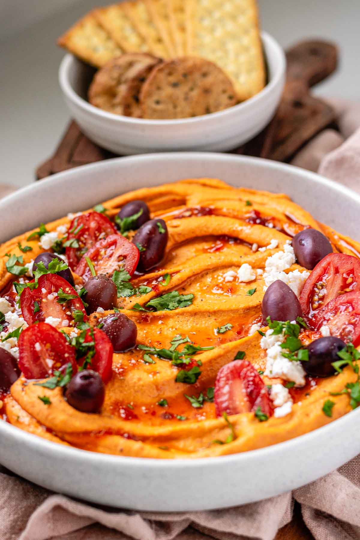 Orange hummus in a bowl topped with olive oil, crumbly white cheese, kalamata olives, chopped herbs, and halved tomatoes beside a bowl of crackers.