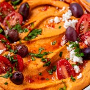 Orange hummus in a bowl topped with olive oil, crumbly white cheese, kalamata olives, chopped herbs, and halved tomatoes.