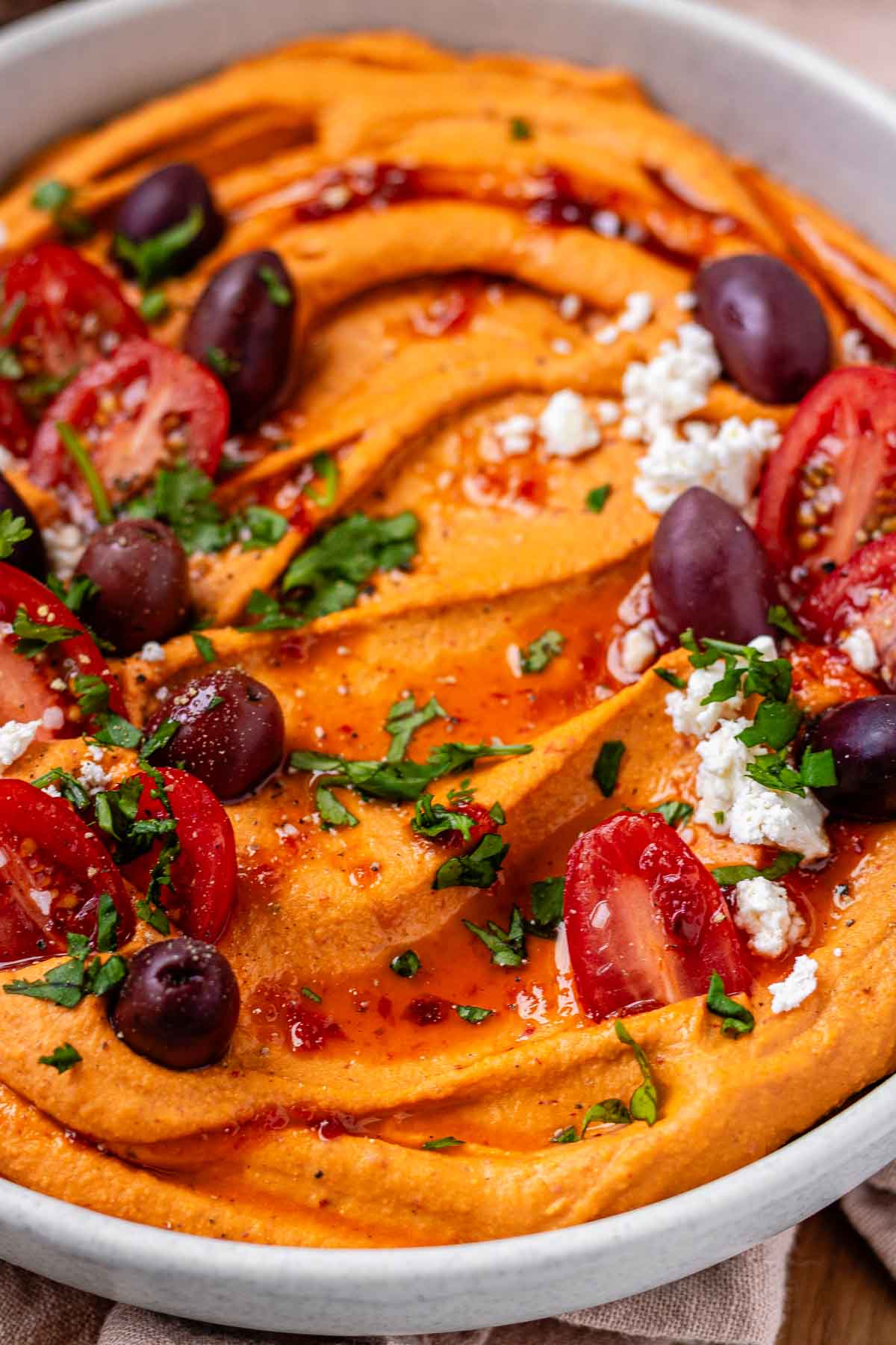 Orange hummus in a bowl topped with olive oil, crumbly white cheese, kalamata olives, chopped herbs, and halved tomatoes.