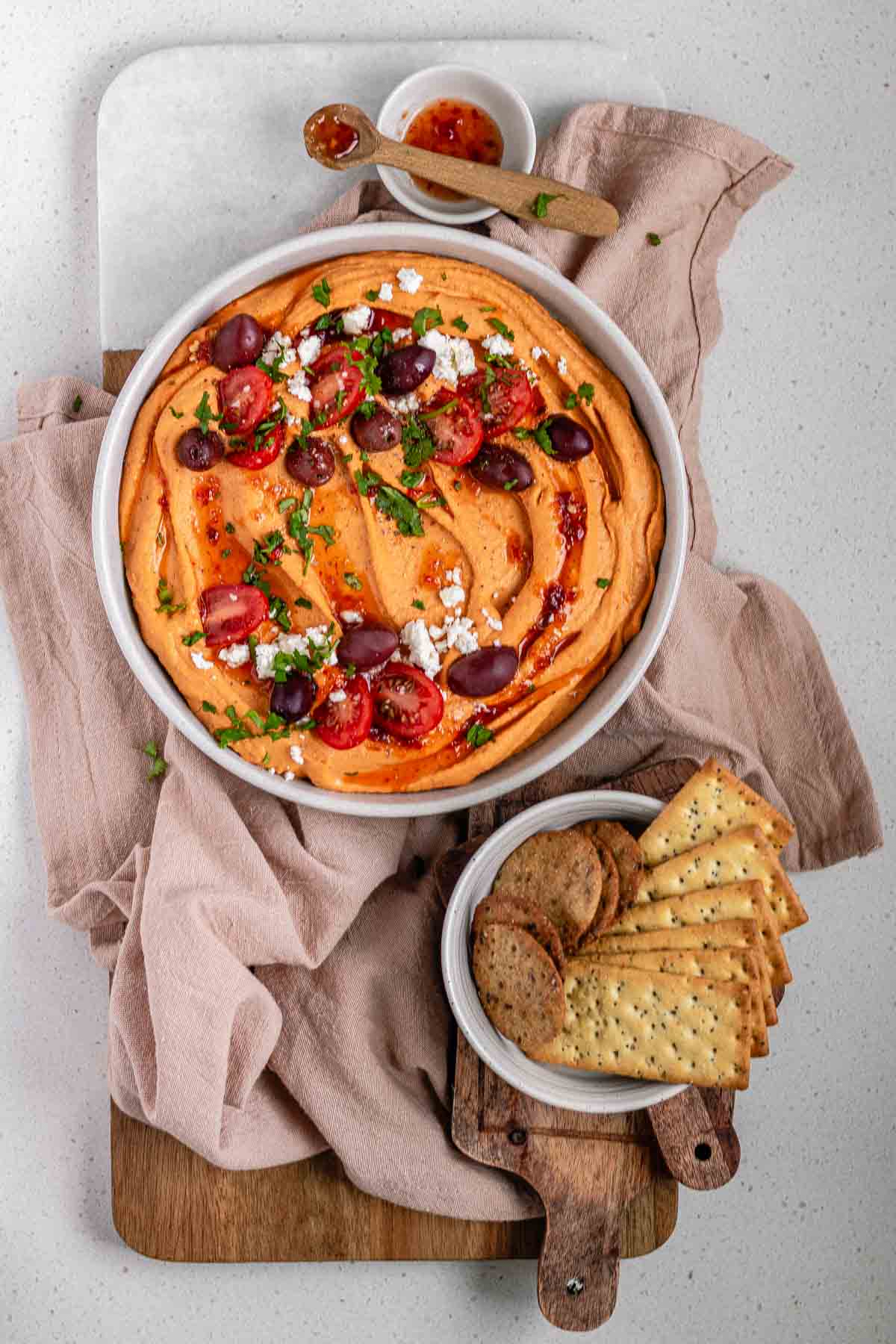 Orange hummus in a bowl topped with olive oil, crumbly white cheese, kalamata olives, chopped herbs, and halved tomatoes beside a bowl of crackers.