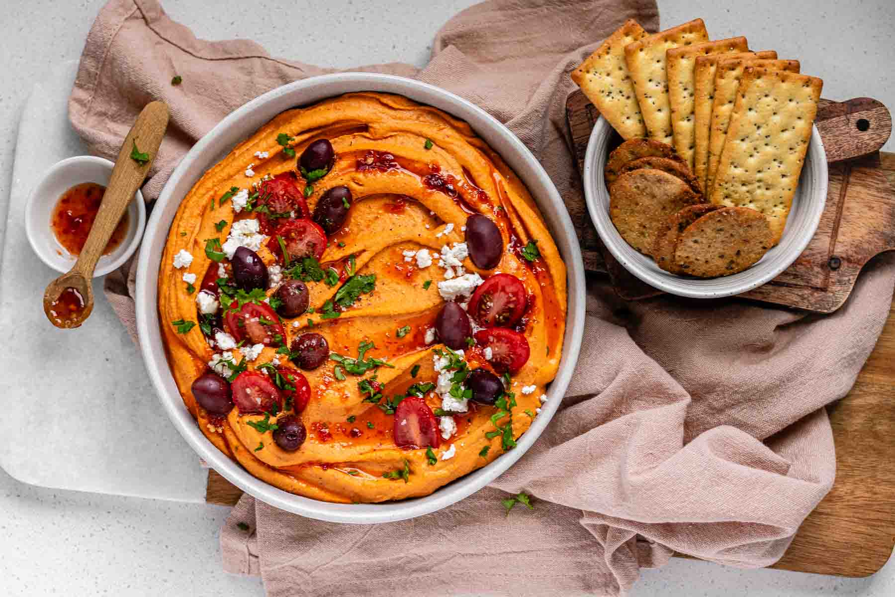 Orange hummus in a bowl topped with olive oil, crumbly white cheese, kalamata olives, chopped herbs, and halved tomatoes beside a bowl of crackers.