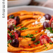 Orange hummus in a bowl topped with olive oil, crumbly white cheese, kalamata olives, chopped herbs, and halved tomatoes with the text hot hummus.
