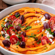 Orange hummus in a bowl topped with olive oil, crumbly white cheese, kalamata olives, chopped herbs, and halved tomatoes with the text hot hummus.