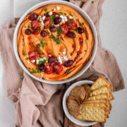 Orange hummus in a bowl topped with olive oil, crumbly white cheese, kalamata olives, chopped herbs, and halved tomatoes beside a bowl of crackers with the text hot hummus.
