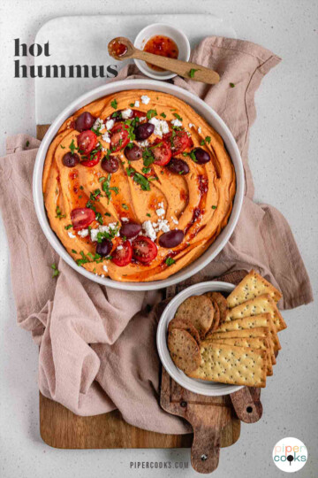 Orange hummus in a bowl topped with olive oil, crumbly white cheese, kalamata olives, chopped herbs, and halved tomatoes beside a bowl of crackers with the text hot hummus.