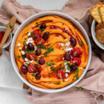 Orange hummus in a bowl topped with olive oil, crumbly white cheese, kalamata olives, chopped herbs, and halved tomatoes.