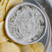 An herby dip in a bowl on a plate with chips.