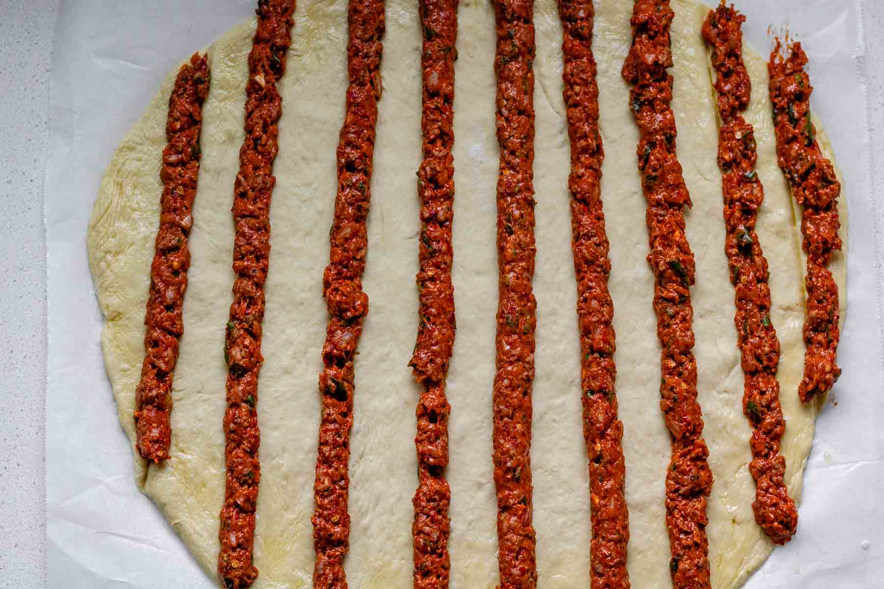 Uncooked flatbread dough topped with lines of uncooked meat.