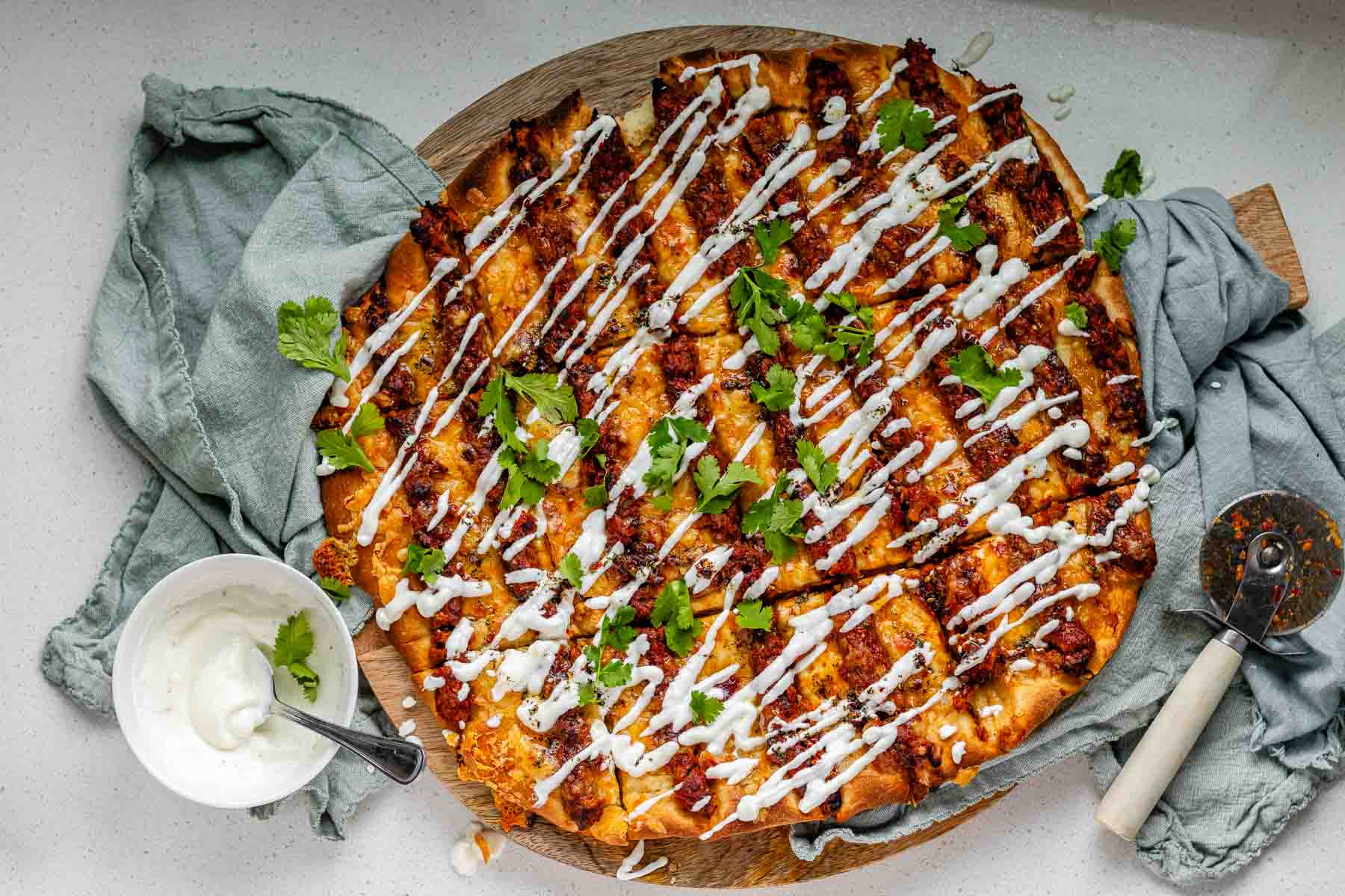 Flatbread topped with lines of cooked meat and cheese, drizzled with yogurt sauce, and scattered with cilantro leaves.