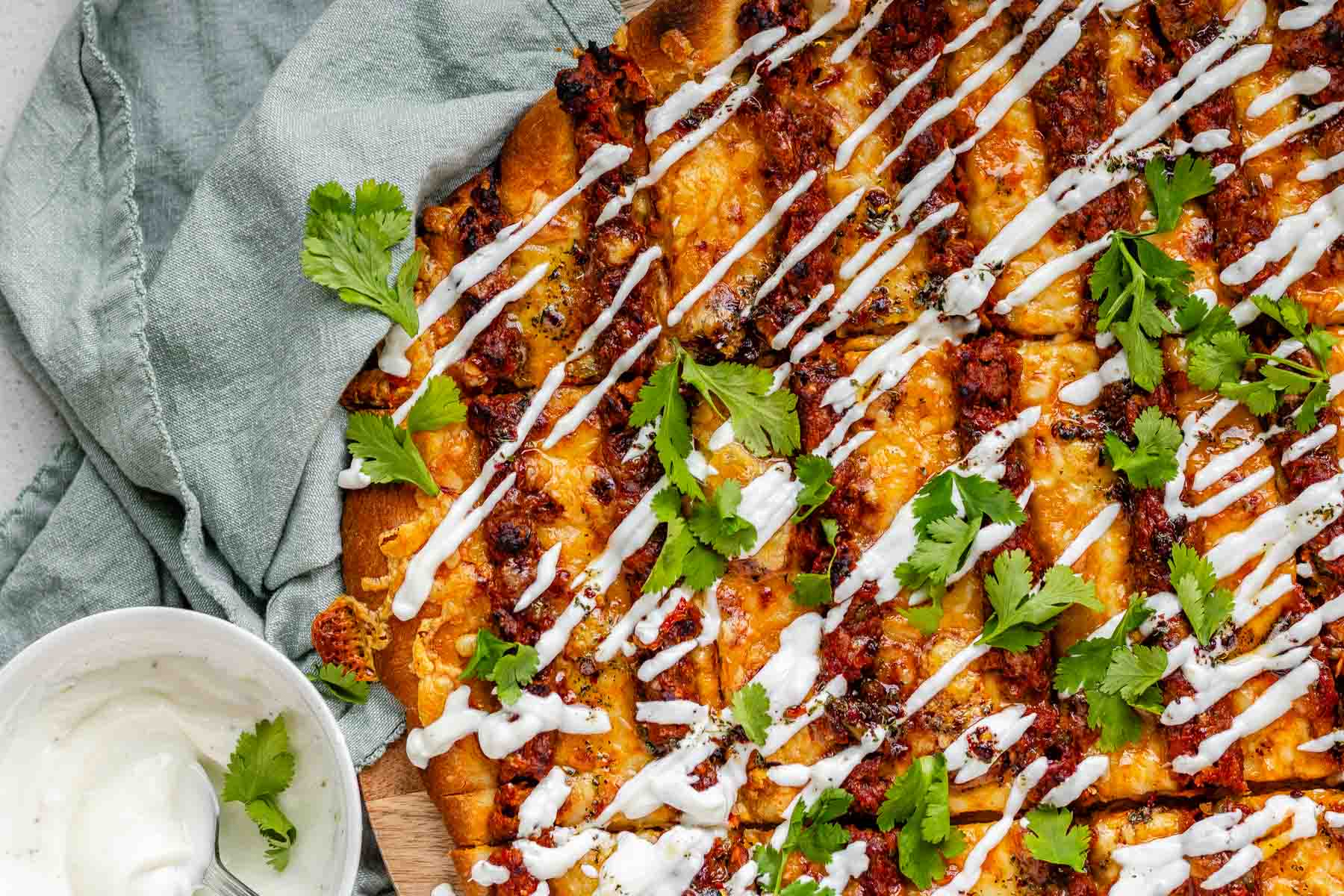 Flatbread topped with lines of cooked meat and cheese, drizzled with yogurt sauce, and scattered with cilantro leaves.