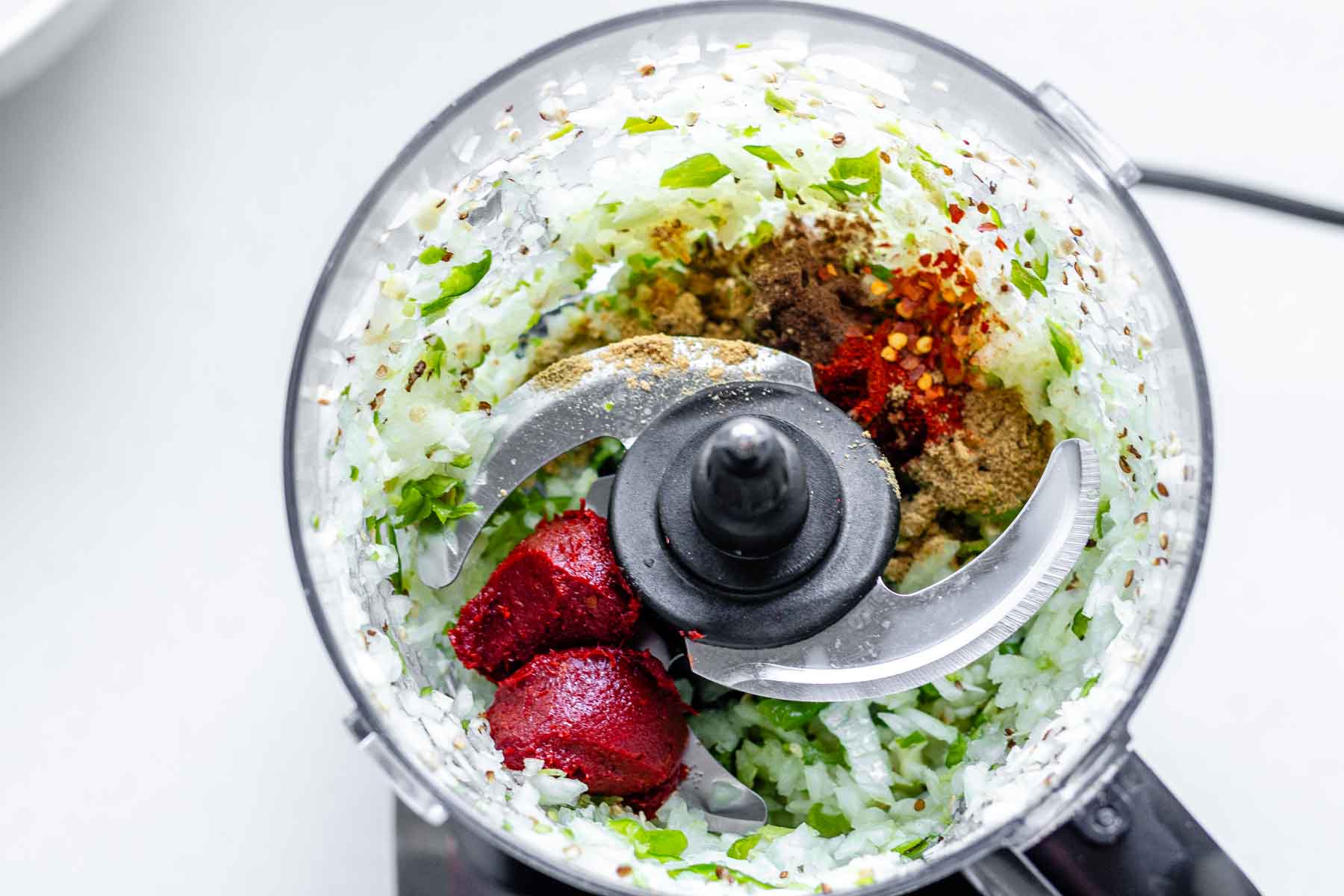 A food processor jar with blended onion, garlic, and green peppers, plus spices and tomato paste.