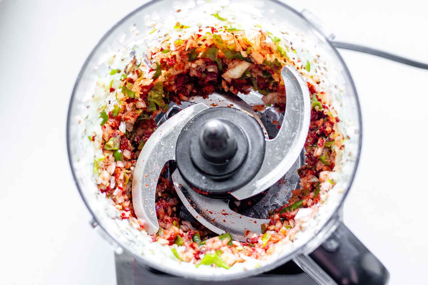 A food processor jar with blended onion, garlic, and green peppers, plus spices and tomato paste.
