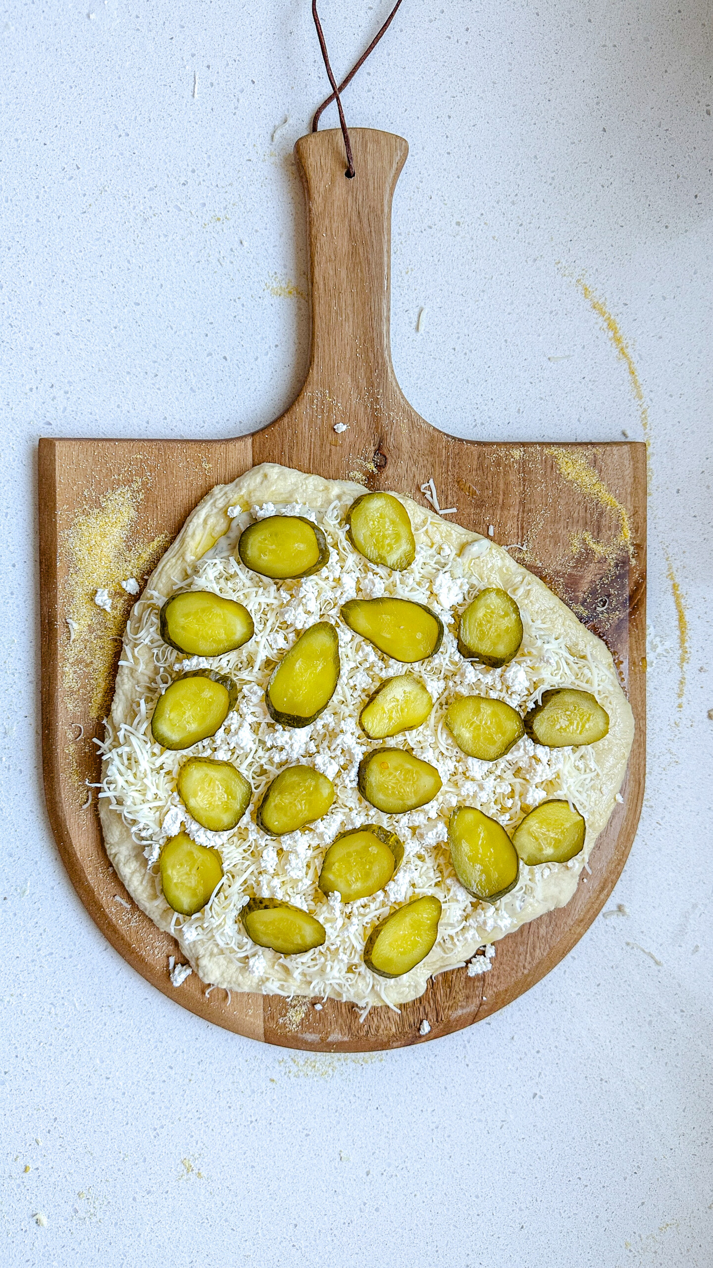 Pizza dough on a pizza peel topped with white sauce, white cheese, and pickles.