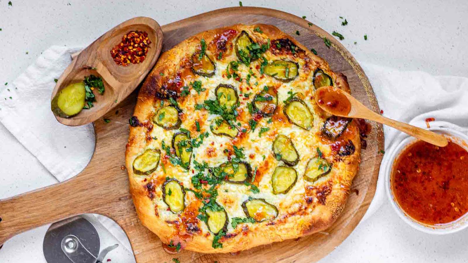 Pizza on a pizza peel topped with pickles and a red sauce.