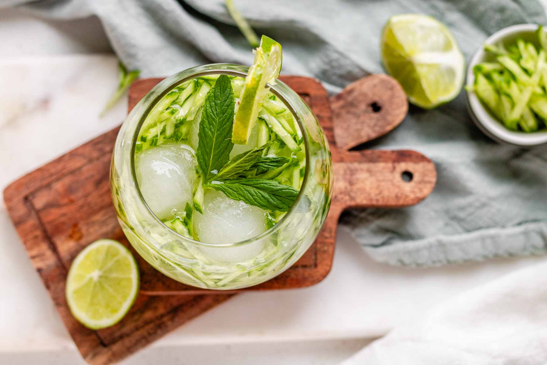 A drink in a glass garnished with julienned cucumber, lime slices, and a sprig of mint.
