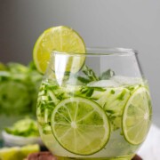 A drink in a glass garnished with julienned cucumber, lime slices, and a sprig of mint.