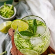 A drink in a glass garnished with julienned cucumber, lime slices, and a sprig of mint.