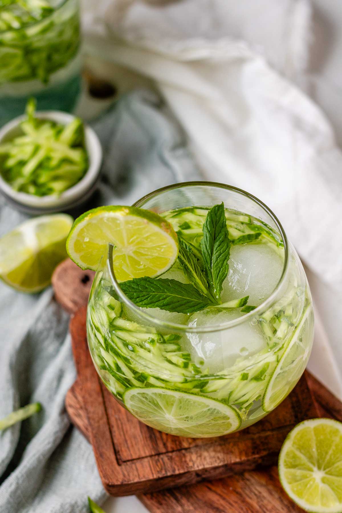 A drink in a glass garnished with julienned cucumber, lime slices, and a sprig of mint.