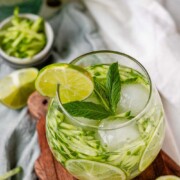 A drink in a glass garnished with julienned cucumber, lime slices, and a sprig of mint with the text sekajabin.